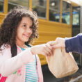 Adult handing brown paper lunch bag to child in front of school bus
