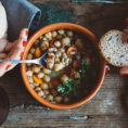 Person using spoon to eat out of a bowl of bean soup with bread in their hand