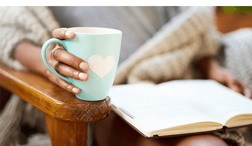 Person holding blue mug while reading a book in a chair
