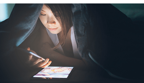 Person looking at digital device under a comforter at night