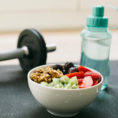Closeup of a bowl of healthy food next to a water bottle and barbell on yoga mat