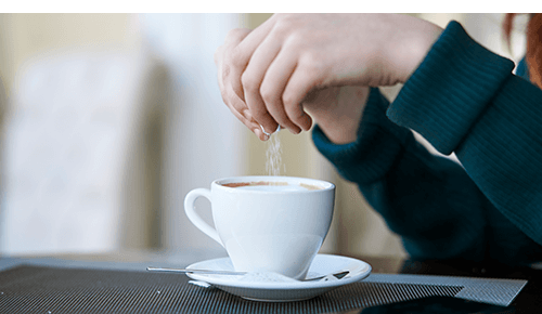 Person putting sugar into cup of coffee sitting on a table Person putting sugar into cup of coffee sitting on a table