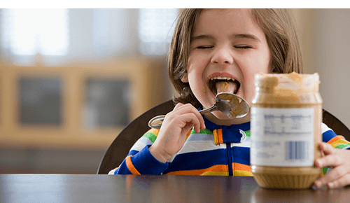 Child licking spoon with peanut butter on it at kitchen counter