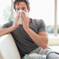 Man sitting on couch blowing his nose with a facial tissue