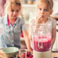 Two children helping make a pink smoothie in a blender at a kitchen counter