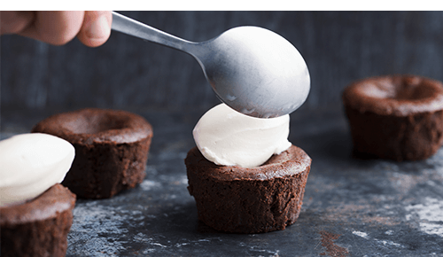Person placing a spoonful of whipped cream on a small chocolate cupcake