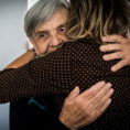 Older smiling woman being embraced by a person with their back to the camera