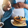 Pregnant person eating a stack of blueberry pancakes with their hand on their stomach