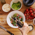 Closeup of person pouring olive oil over bowl of salad on kitchen counter surrounded by other fresh vegetables