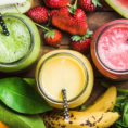 Birds-eye view of three different fruit smoothies in glass cups surrounded by a variety of fresh fruit