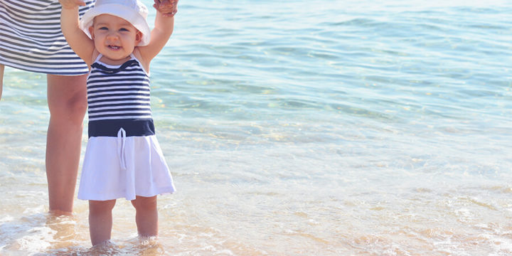 Infant walking with adult help on a beach near a body of water