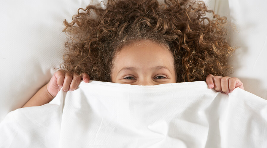 Young child smiling with bed sheets pulled up to their eyes