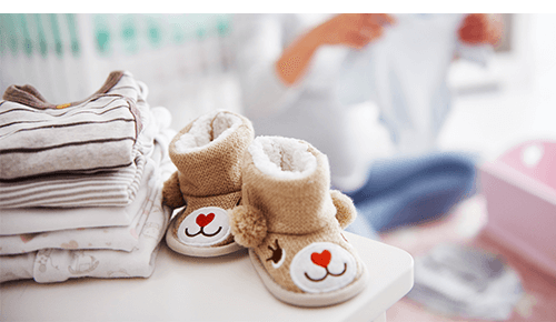 Baby shoes and clothing sitting on dresser with pregnant person looking through clothing in the background Baby shoes and clothing sitting on dresser with pregnant person looking through clothing in the background