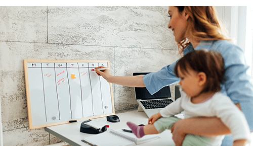 Woman holding infant while writing on a whiteboard with days of the week written on it Woman holding infant while writing on a whiteboard with days of the week written on it