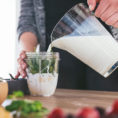 Person pouring milk into a smoothie blender glass that has spinach and fruit in it