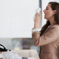 Person sneezing into facial tissue while seated at work desk