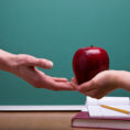 Child handing red apple to adult teacher in front of classroom chalkboard