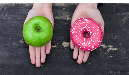 Two hands, one holding a green apple and one holding a pink frosted donut