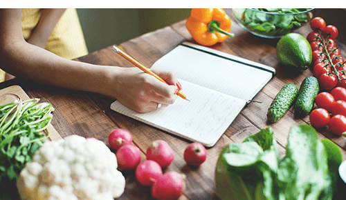 Person writing in journal on table covered with a variety of vegetables