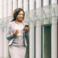 Woman in business work clothes walking while looking at her mobile phone and holding a coffee cup