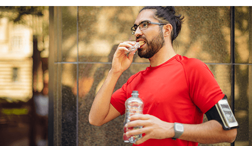 Person eating protein bar while walking outside and holding water bottle after exercising