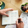 Person writing in diet and workout journal with a salad on the counter next to them