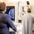 Clinical staff viewing screen as woman in background receives a mammogram