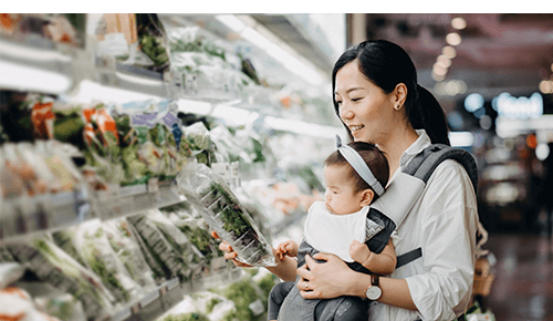 Mother with a baby in a carrier looking a lettuce package at the grocery store