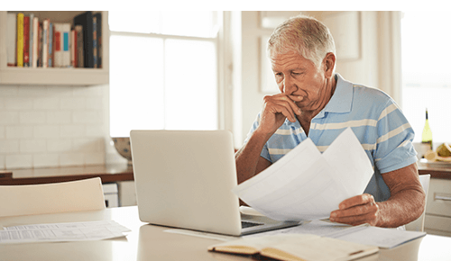 Man at kitchen counter looking at computer while holding papers