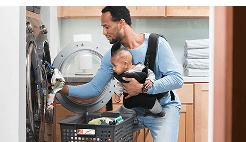 Dad putting laundry in a washing machine while holding baby in carrier