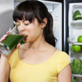 Person cringing as they drink a healthy green drink in front a fridge full of vegetables and fruit