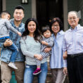 Three generations of an Asian family smiling and standing in front of house