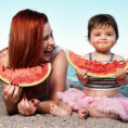 woman and child lay on beach eating watermelon and laughing